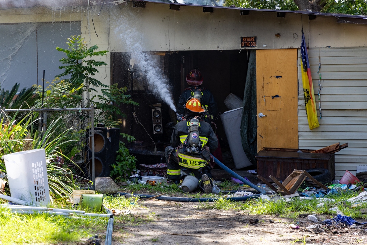 Two firefighters spray water into a smoke-damaged home in Ocala surrounded by debris and overgrown vegetation.