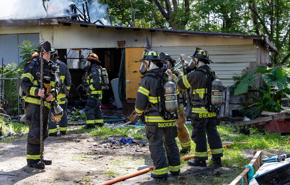 Two firefighters in gear stand near the smoky exterior of a single-story house with boarded windows.