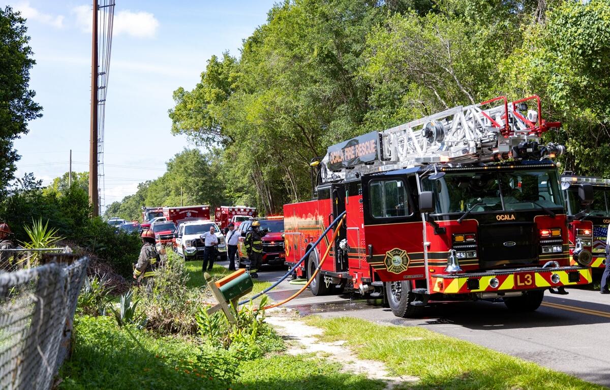 Multiple fire trucks and emergency vehicles parked along a road lined with trees while firefighters respond to a house fire.