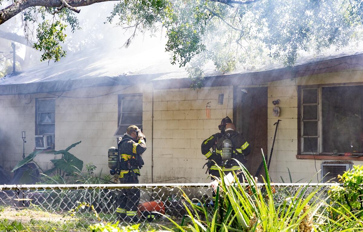 Two firefighters in gear stand near the smoky exterior of a single-story house with boarded windows.