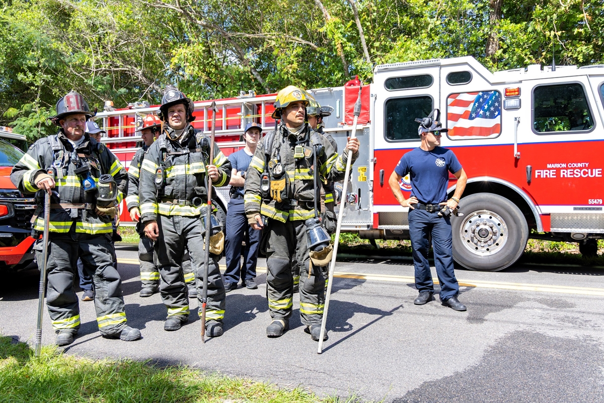 A group of firefighters, some holding tools, stand in front of a fire engine after battling a house fire.