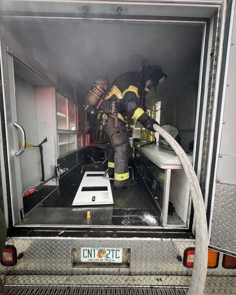 A firefighter enters the back of a heavily smoked ambulance with a hose, as cabinet doors lie open and scorched inside.