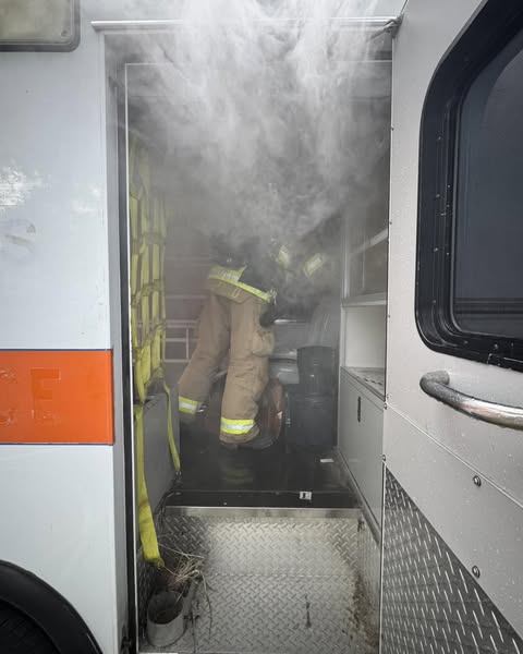 Thick smoke pours out of an ambulance as a firefighter in full gear kneels inside, addressing the source of the fire.