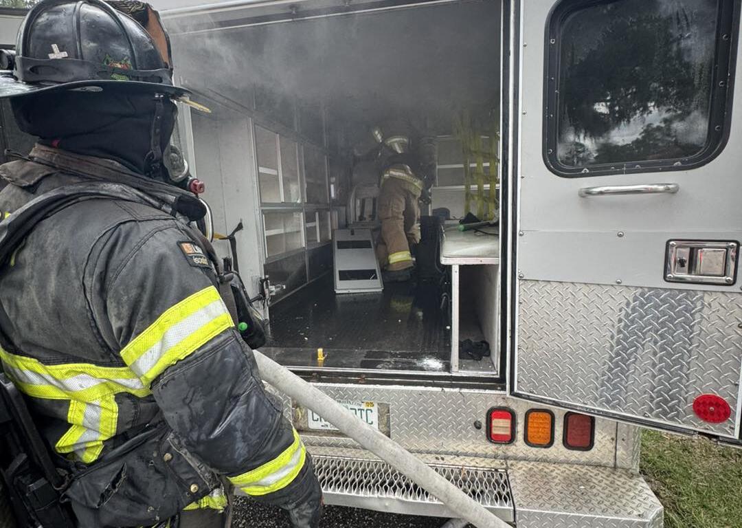 A firefighter stands outside the rear of a smoke-filled ambulance, holding a hose while another firefighter works inside.