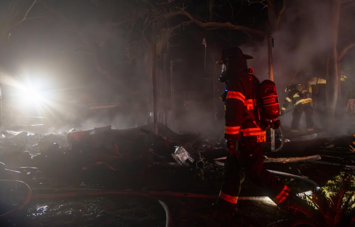 Firefighters are seen actively working at the scene of a brush or structural fire near a wooded area. Smoke is visible in the air, and fire hoses are laid out across the ground.