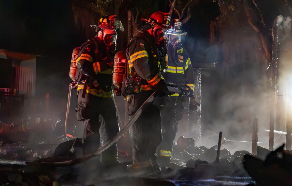 This image captures a firefighter in full gear walking toward a structure that has sustained severe fire damage. The roof appears partially collapsed and charred, and remnants of the building are still smoking.