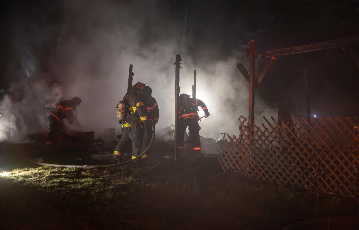 A close-up view of a collapsed and burned-out structure. Debris is scattered around, with heavy smoke or steam still emanating from the ruins. The scene suggests the fire has recently been extinguished, and firefighters are likely in the overhaul phase, checking for hotspots.