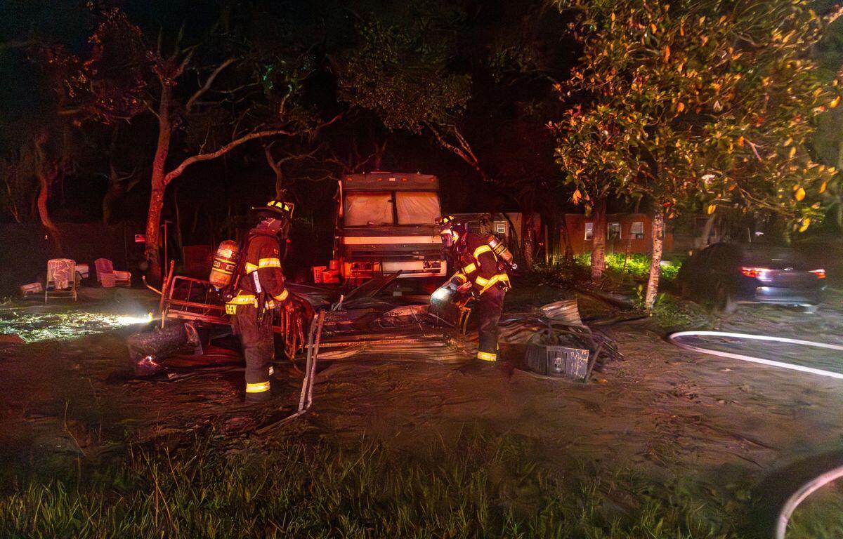 In this nighttime scene illuminated by fire truck lights and spotlights, firefighters in full gear sift through charred debris in front of a heavily damaged mobile home. The area is surrounded by trees and nearby vehicles, with a garden hose lying on the ground.