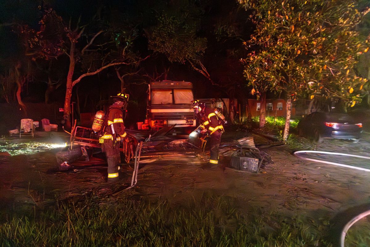 In this nighttime scene illuminated by fire truck lights and spotlights, firefighters in full gear sift through charred debris in front of a heavily damaged mobile home. The area is surrounded by trees and nearby vehicles, with a garden hose lying on the ground.