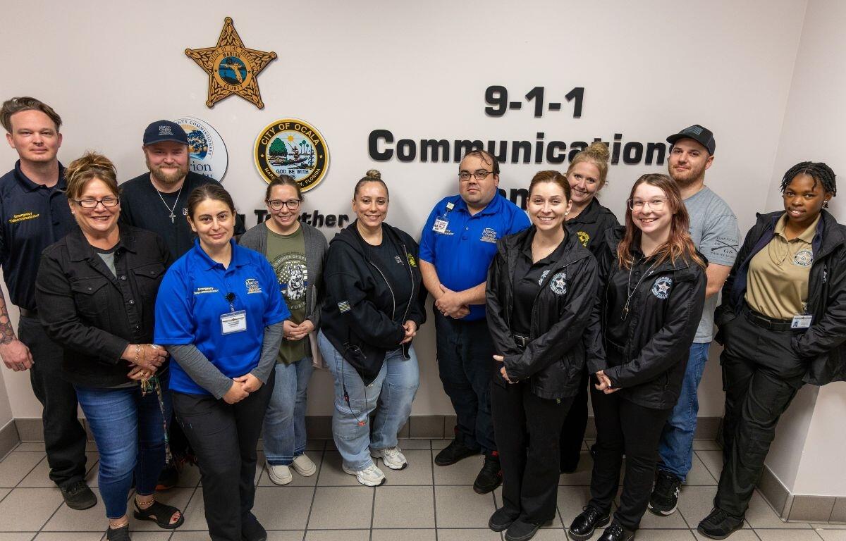 Group of Marion County 911 dispatchers and staff standing in front of wall with agency logos at communications center.