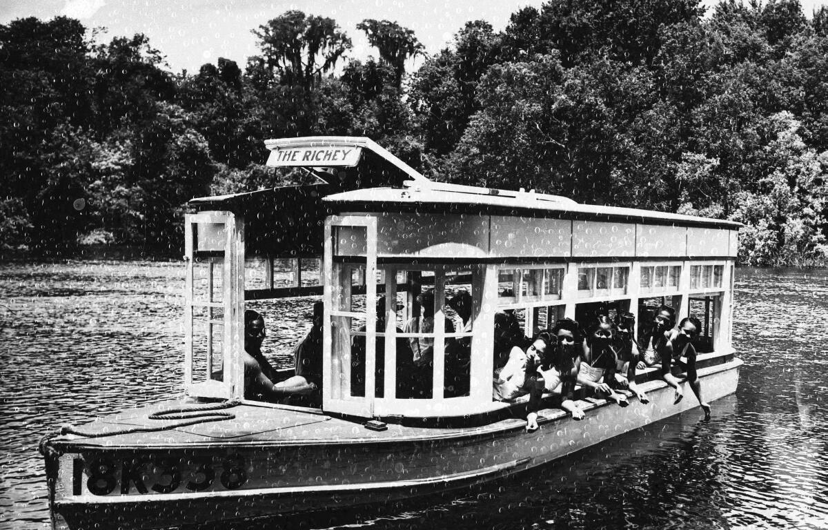 A group of African American women aboard a boat signed, "The Richey." The women are smiling, waving as the boat floats on the clear spring waters surrounded by trees and vegetation. 