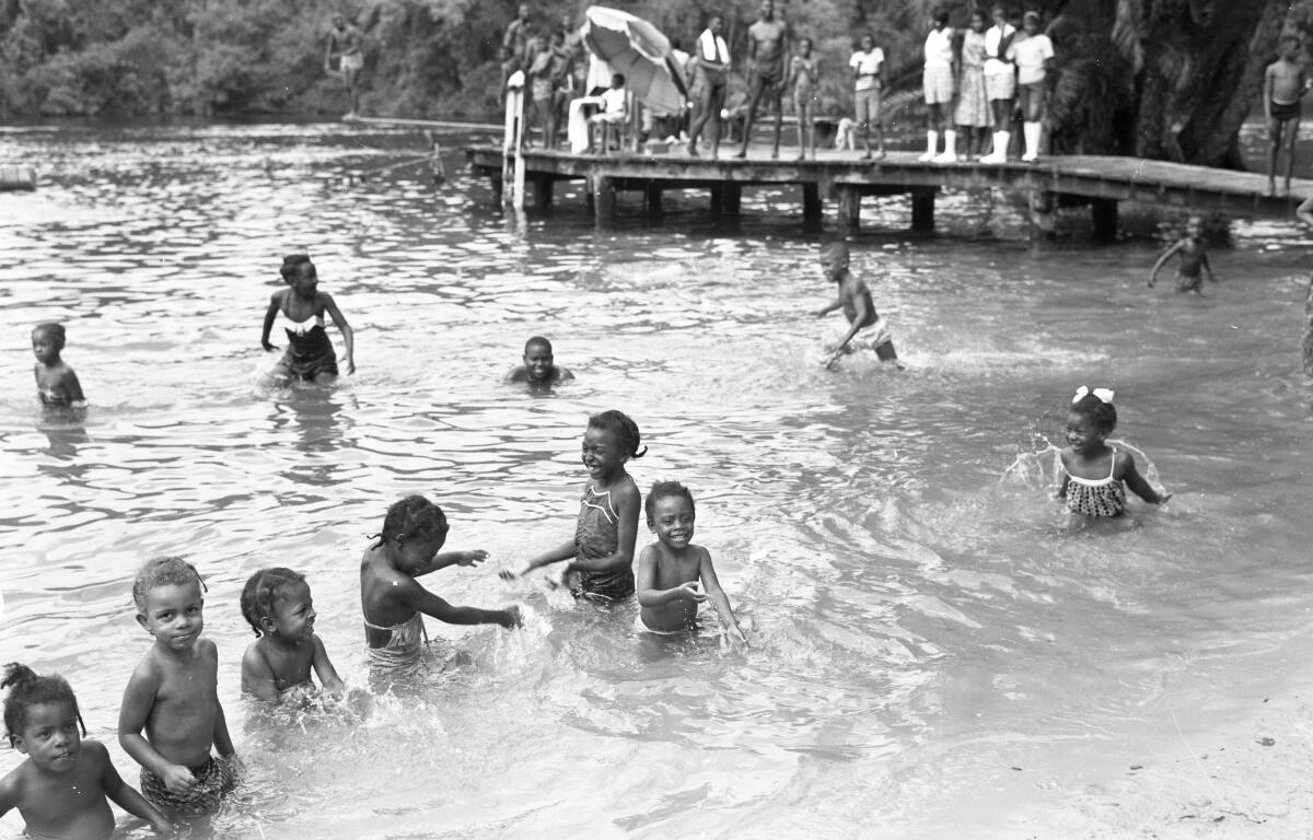 African American children playing in shallow water and on a sandy beach area. The kids are splashing and wading, with trees and a dock in the background.