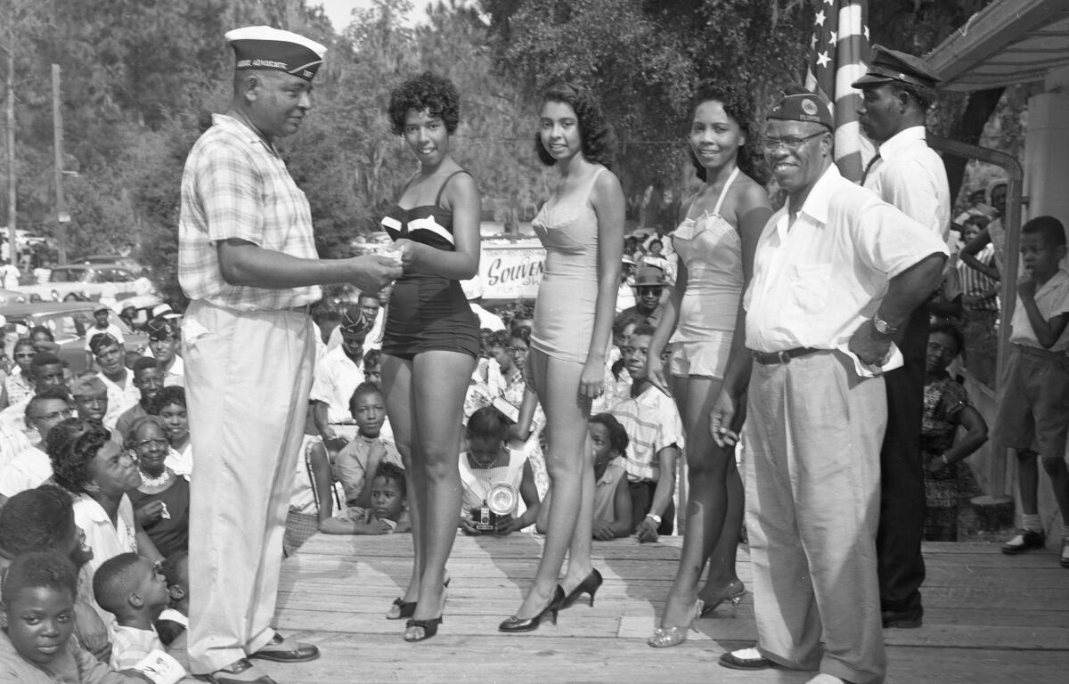 An African American women receiving a trophy for winning a beauty pageant at Paradise Park. She stands smiling in a dress as a man presents the award, with a crowd of onlookers in the background.