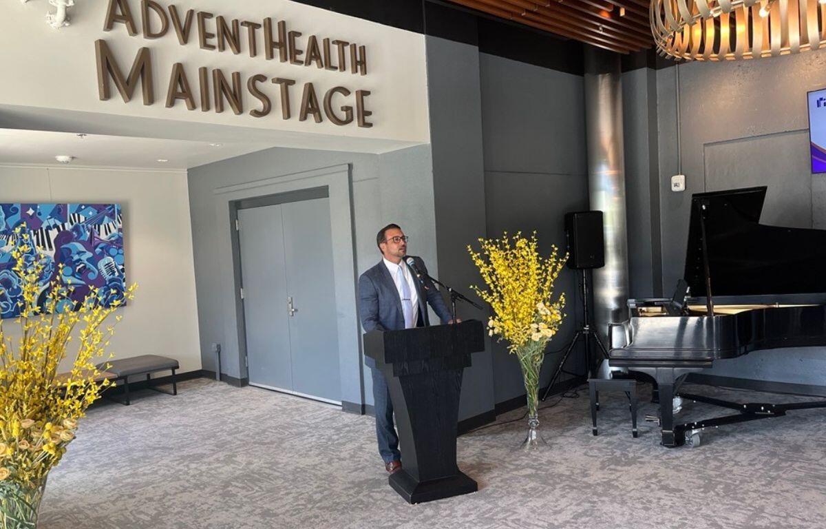 A man dressed in a suit, standing behind a podium with a microphone, with a wall with lettering to the man's left, with ornamental flora, a chair and gray wall to his right.