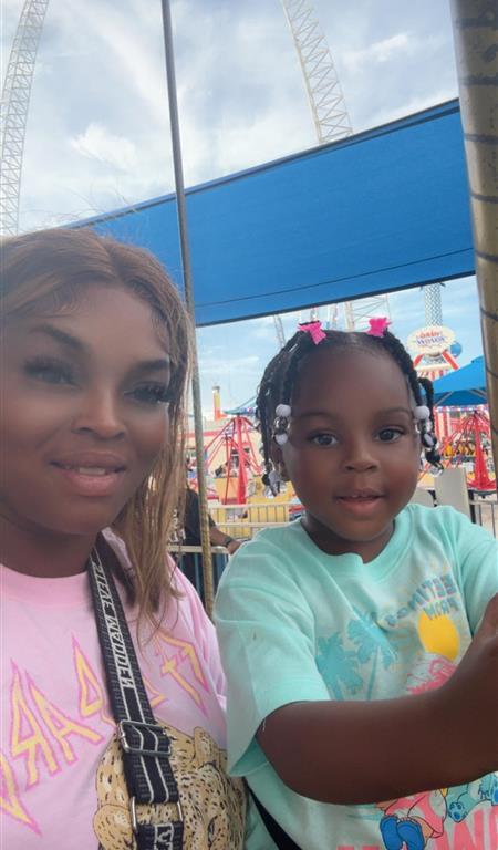 A smiling woman and a young girl enjoying time together at an amusement park, with colorful rides and a blue canopy in the background.