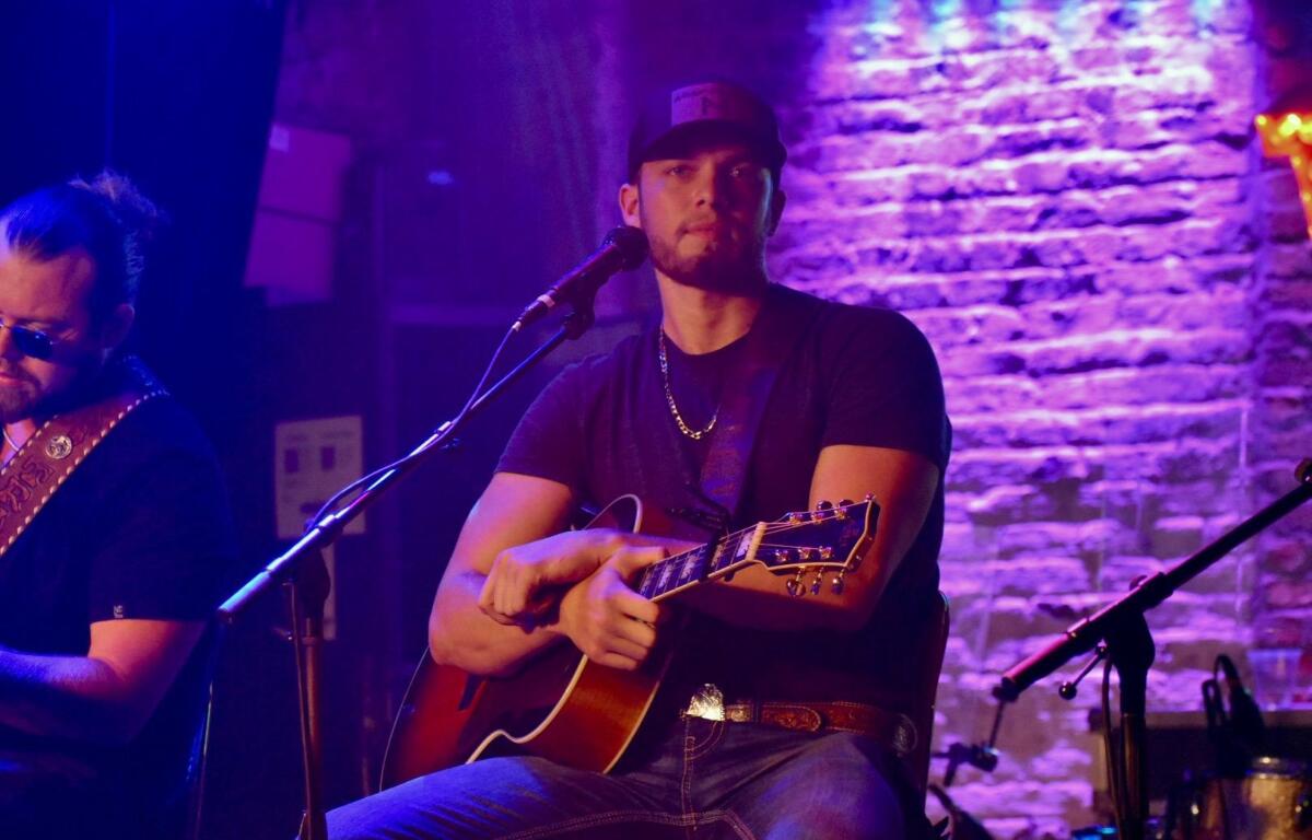 A musician in a black shirt and cap plays acoustic guitar under vibrant purple stage lights during a live performance.