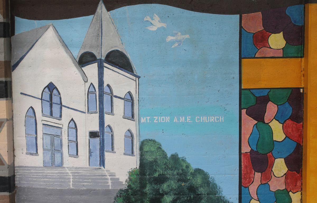 A white church with a gray steeple and blue windows, with a blue sky in the background, with white birds and green trees to the right of the church.