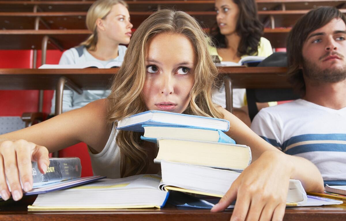 Student overwhelmed by textbooks in a lecture hall while others talk in the background.