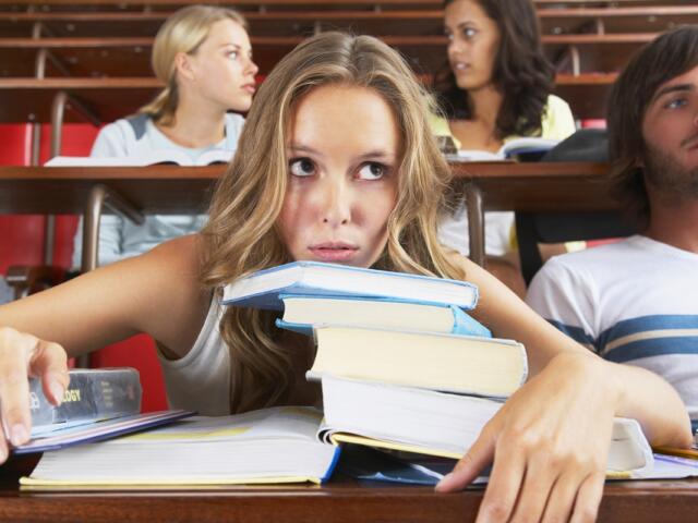 Student overwhelmed by textbooks in a lecture hall while others talk in the background.