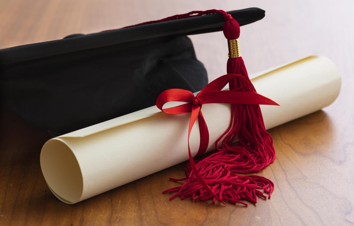 Graduation cap with red tassel and rolled diploma tied with red ribbon on a wooden table.