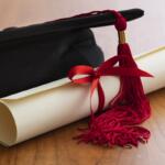 Graduation cap with red tassel and rolled diploma tied with red ribbon on a wooden table.