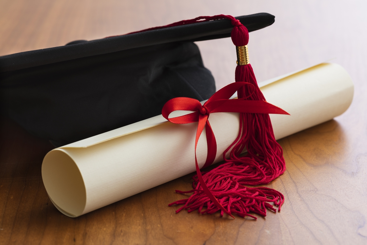 Graduation cap with red tassel and rolled diploma tied with red ribbon on a wooden table.