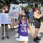 A group of demonstrators hold protest signs during the No Kings rally on Ocala's Downtown Square, including a woman in front wearing a purple "No Kings Since 1776" shirt.
