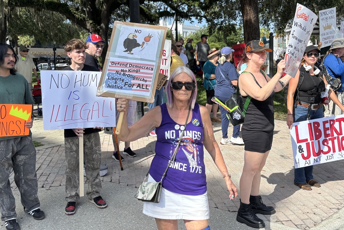 A group of demonstrators hold protest signs during the No Kings rally on Ocala's Downtown Square, including a woman in front wearing a purple "No Kings Since 1776" shirt.