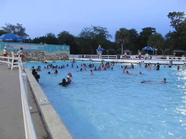 A large outdoor swimming pool filled with people enjoying the water during the evening. Lifeguards are present around the pool deck, and there are umbrellas and signage in the background, including one that says 'Wild Waters.' Trees and palm trees surround the pool area.