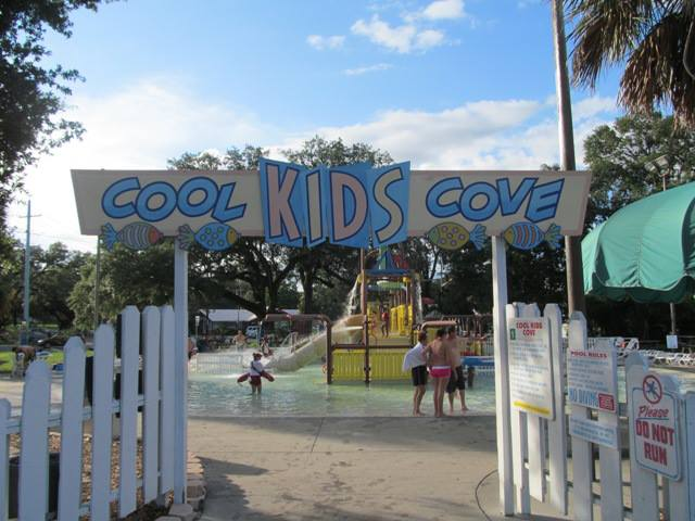Entrance to a children’s splash area named “Cool Kids Cove,” featuring a colorful sign with playful lettering above a gated white picket fence. Inside, kids are playing in shallow water with small slides and water features, surrounded by trees and shaded structures. A sign on the right reads "Please Do Not Run." The atmosphere is bright and sunny.