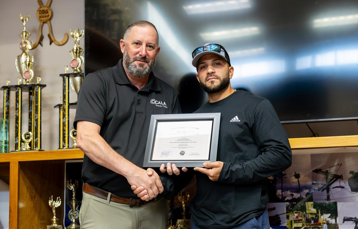 Gabriel Just-Rodriguez holds his framed apprenticeship certificate and shakes hands with a supervisor at the Ocala Utility Services graduation ceremony.
