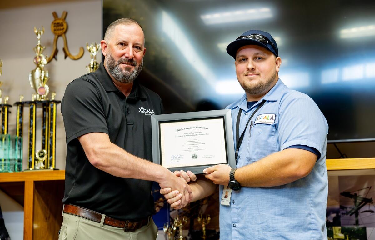 Chris Kennedy shakes hands with a supervisor while holding his framed certificate at the Ocala Utility Services apprenticeship graduation ceremony.