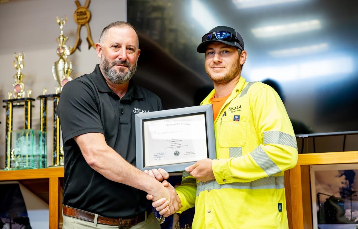 Ocala Electric Utility Jeremy Retherford receives a framed apprenticeship certificate and shakes hands with a supervisor at the June 2025 graduation ceremony.