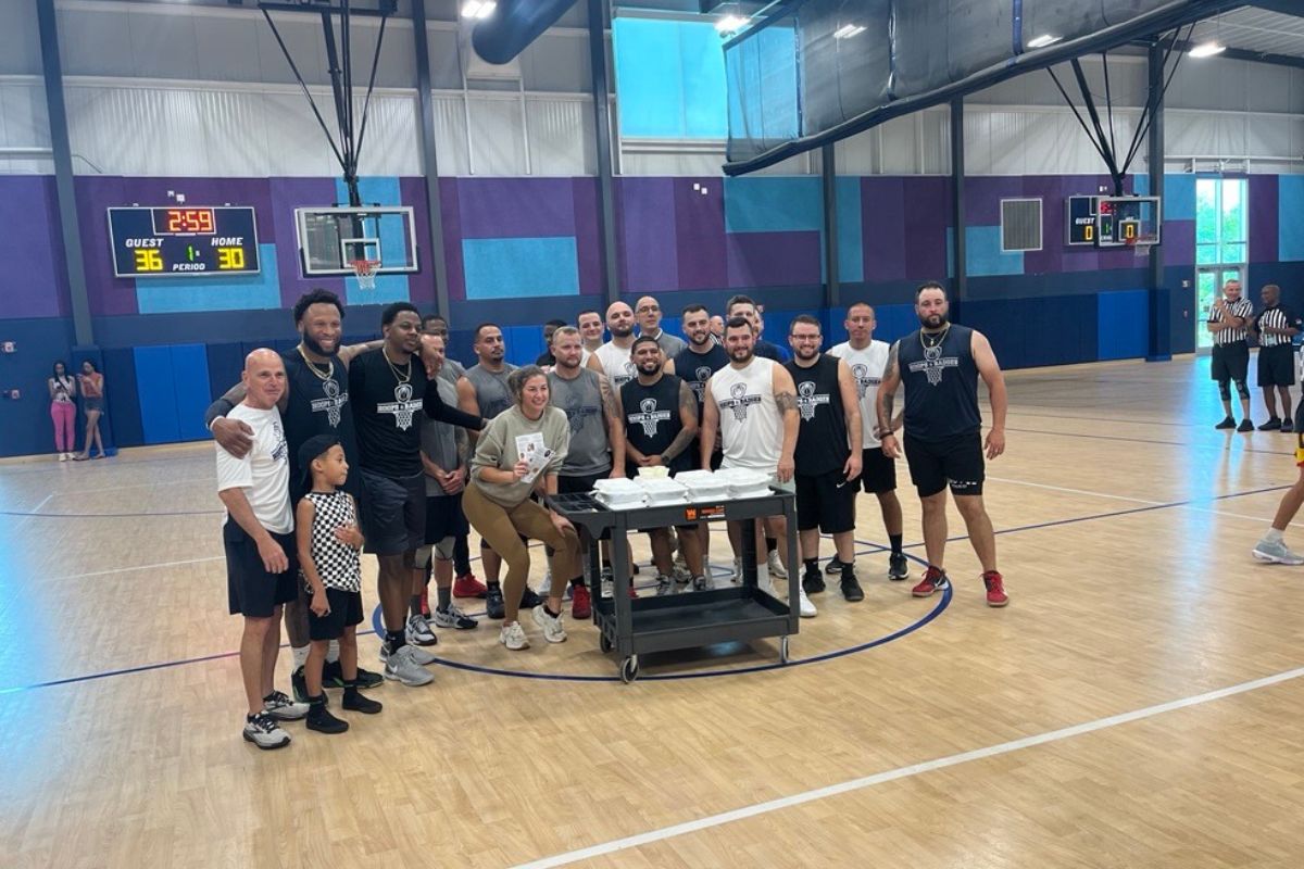 A gym floor in the foreground with a table full of refreshments in front of a group of men, with a blue and white wall in the background. 