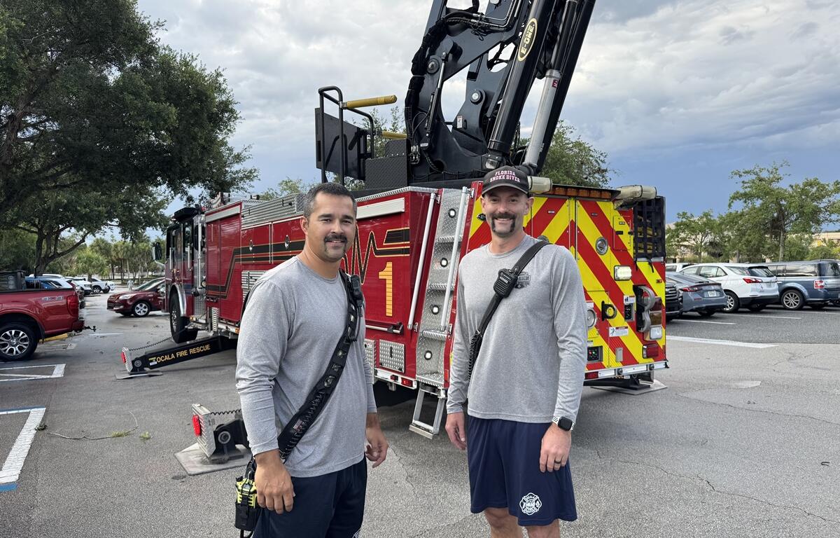 Two Ocala firefighters in gray shirts and navy shorts stand in front of a fire truck at a PTSD Awareness Walk event.