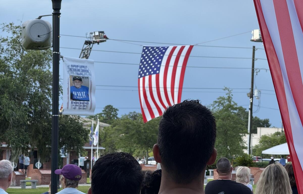 Participants stand for the national anthem as a large American flag is displayed overhead during the PTSD Awareness Walk in Ocala, Fla., on Friday, June 27, 2025. The flag was raised by Ocala Fire Rescue to honor veterans and first responders.