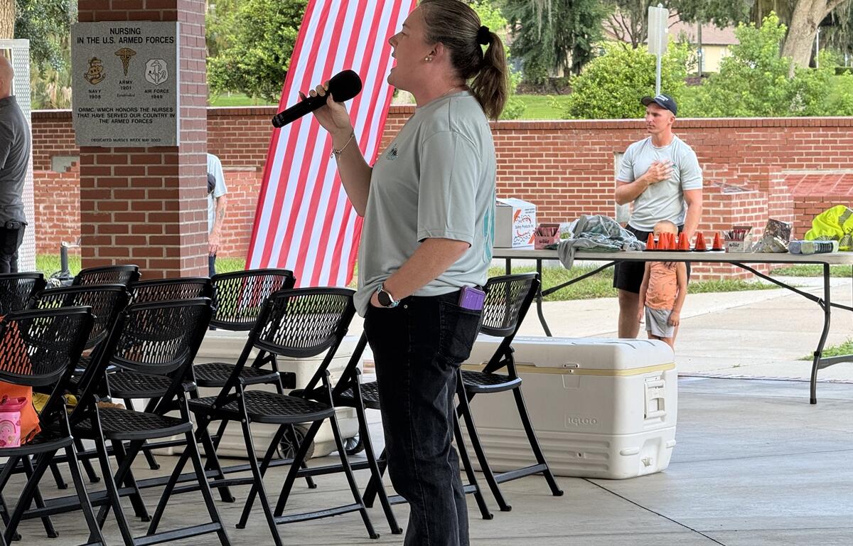 Woman sings into a microphone while others stand respectfully during the national anthem at the PTSD Awareness Walk.