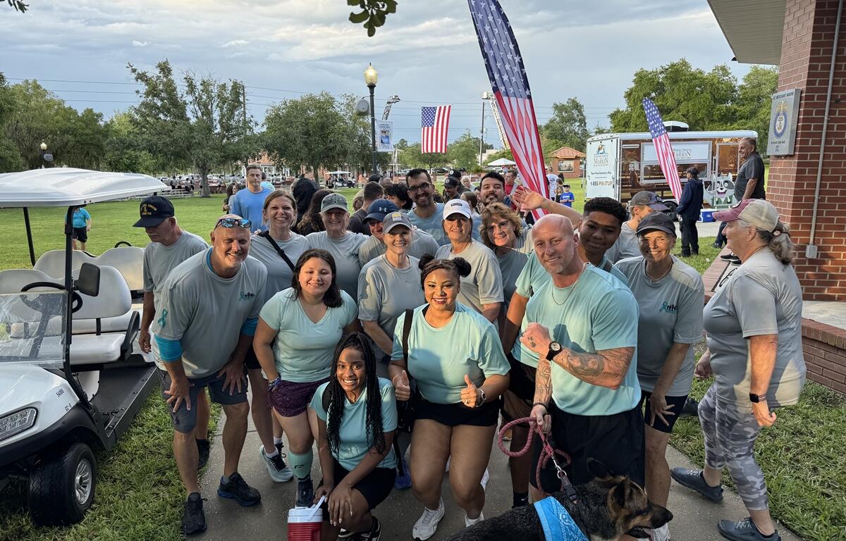 Smiling group of PTSD Awareness Walk participants pose together near the starting area with flags and a service dog.