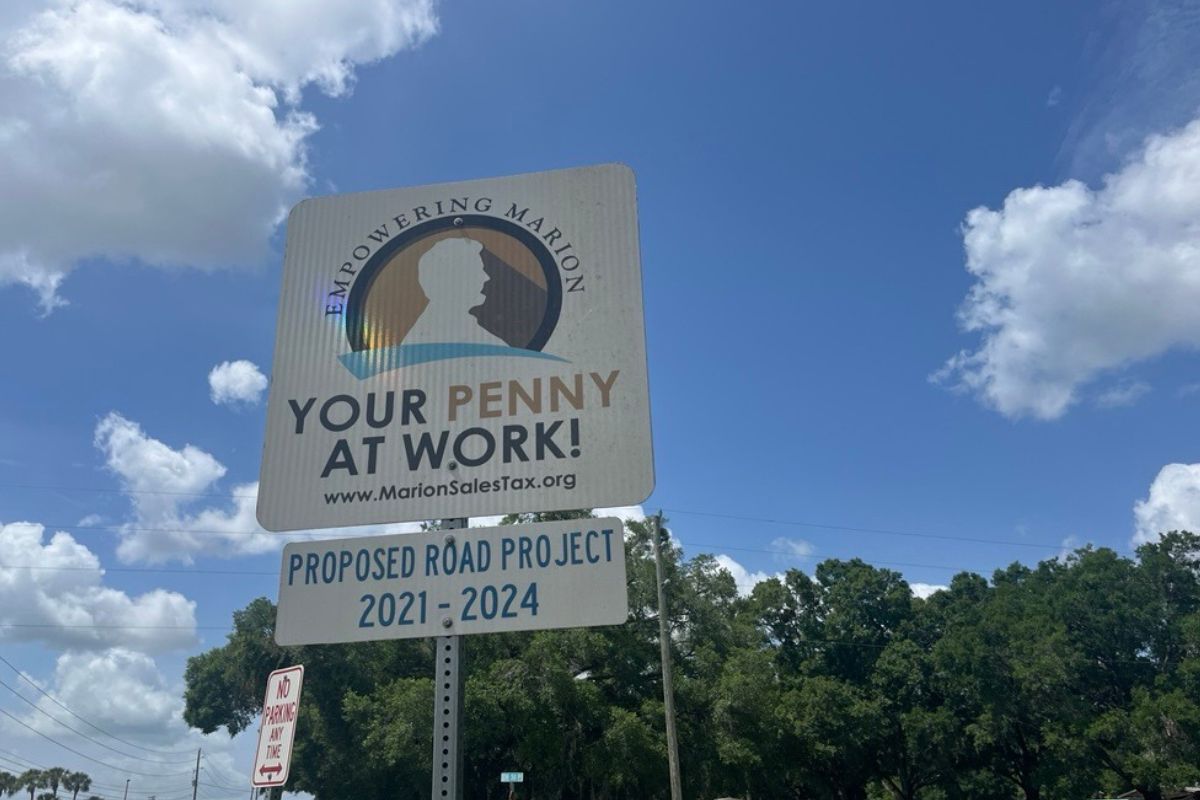 A white placard sign on top of a white rectangular sign, with green trees and blue sky with white clouds in the background. 