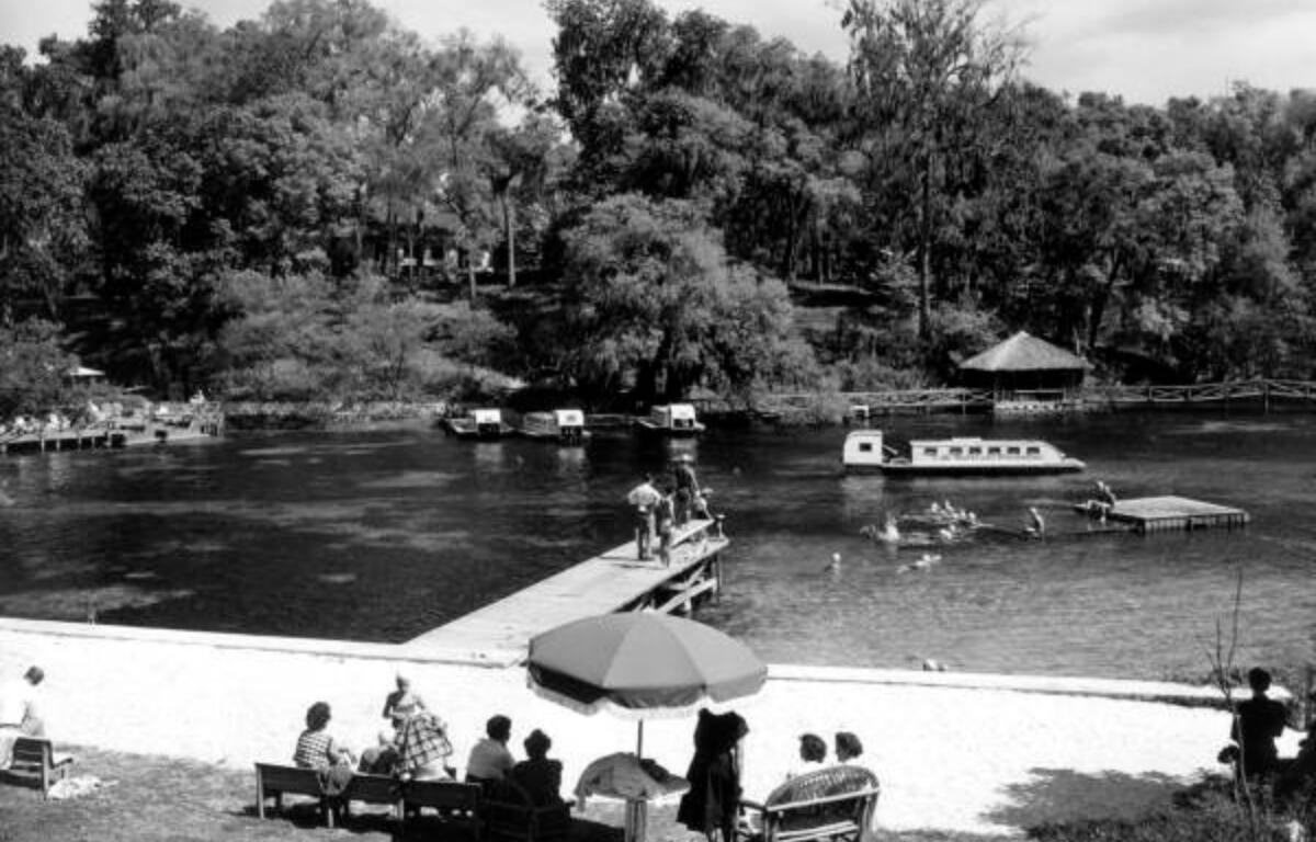 A black-and-white photo of a riverside recreation area shows people gathered on benches along a sandy shoreline, with a large umbrella in the center foreground. A wooden dock extends into the water, where several people are swimming and interacting. Multiple covered tour boats are seen on the river, and dense trees line the opposite bank. A thatched-roof hut is partially visible among the trees. The setting suggests a popular leisure spot, likely from the mid-20th century.