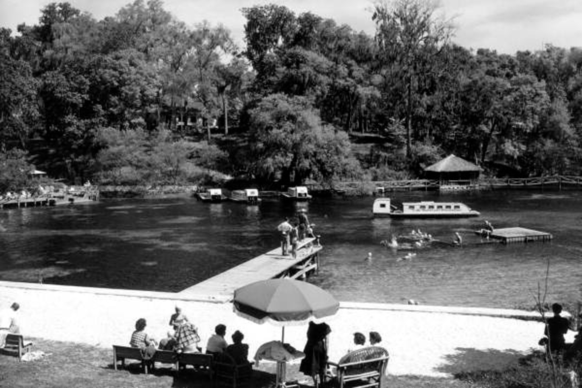 A black-and-white photo of a riverside recreation area shows people gathered on benches along a sandy shoreline, with a large umbrella in the center foreground. A wooden dock extends into the water, where several people are swimming and interacting. Multiple covered tour boats are seen on the river, and dense trees line the opposite bank. A thatched-roof hut is partially visible among the trees. The setting suggests a popular leisure spot, likely from the mid-20th century.