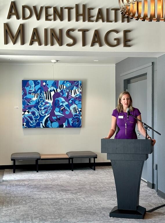 Natalie McComb speaking at a podium beneath the AdventHealth Mainstage sign at the Reilly Arts Center.