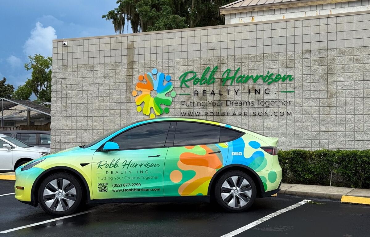A brightly wrapped Robb Harrison Realty vehicle is parked in front of the company’s new Ocala office, featuring a colorful logo and branding on the building’s exterior wall.