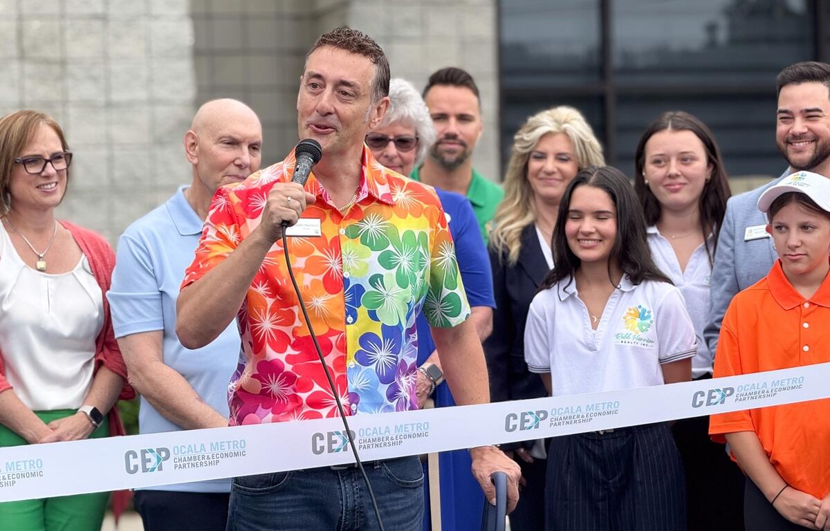 Robb Harrison speaks into a microphone at his realty firm's ribbon-cutting event, standing in front of a crowd behind a CEP-branded ribbon outside the new Ocala office.