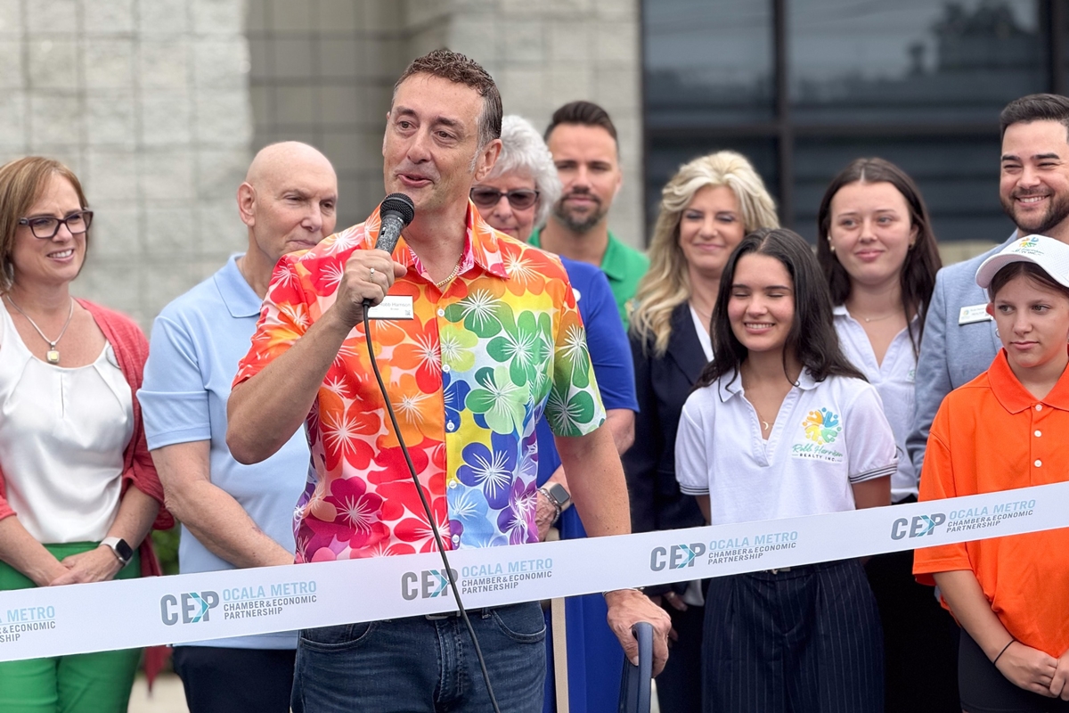 Robb Harrison speaks into a microphone at his realty firm's ribbon-cutting event, standing in front of a crowd behind a CEP-branded ribbon outside the new Ocala office.