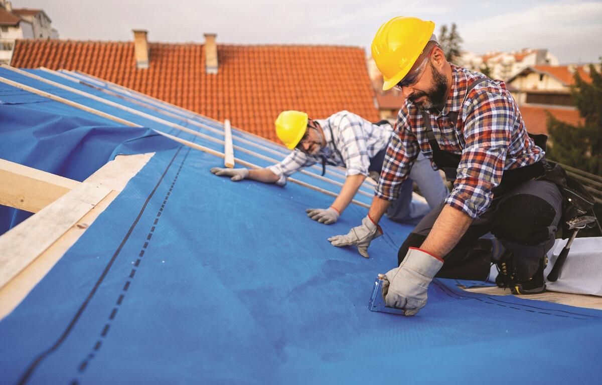 Two roofers in hard hats install blue underlayment on a house roof, using staple guns and wooden beams.