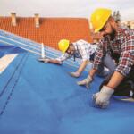 Two roofers in hard hats install blue underlayment on a house roof, using staple guns and wooden beams.