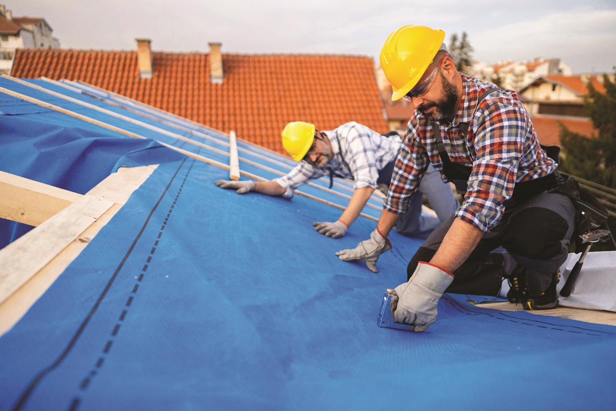 Two roofers in hard hats install blue underlayment on a house roof, using staple guns and wooden beams.