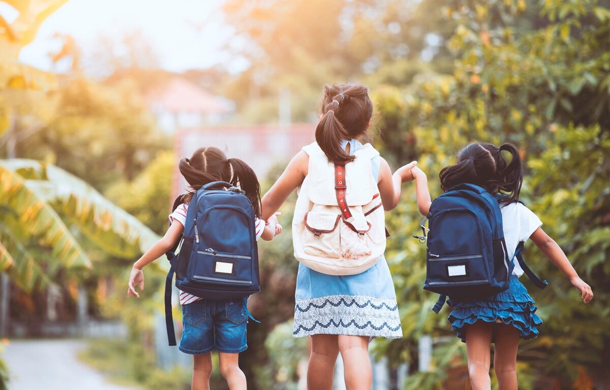 Three young girls wearing backpacks walk hand-in-hand down a tree-lined path.