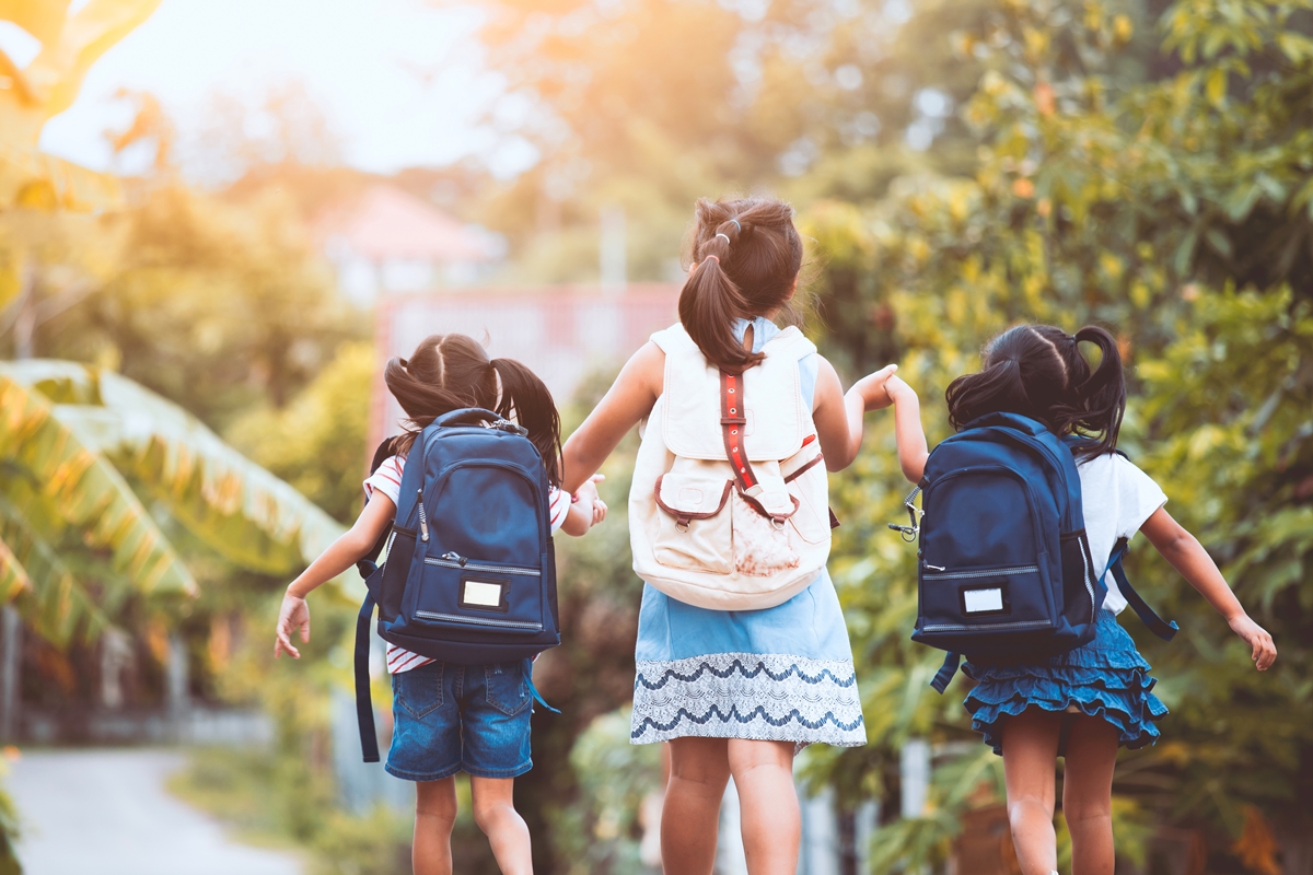 Three young girls wearing backpacks walk hand-in-hand down a tree-lined path.